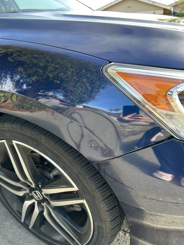 A dent in the front fender of a dark blue sedan, just above the wheel
